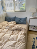 Unmade bed with beige sheets and blue pillows in a room with a window and nightstand.