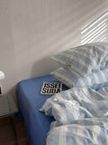 Bed with striped bedding and a pillow, featuring a 'ISSEI SUDA' sign on a white wall.