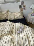 Bed with striped bedding and pillows, with a small table and lamp in the background.