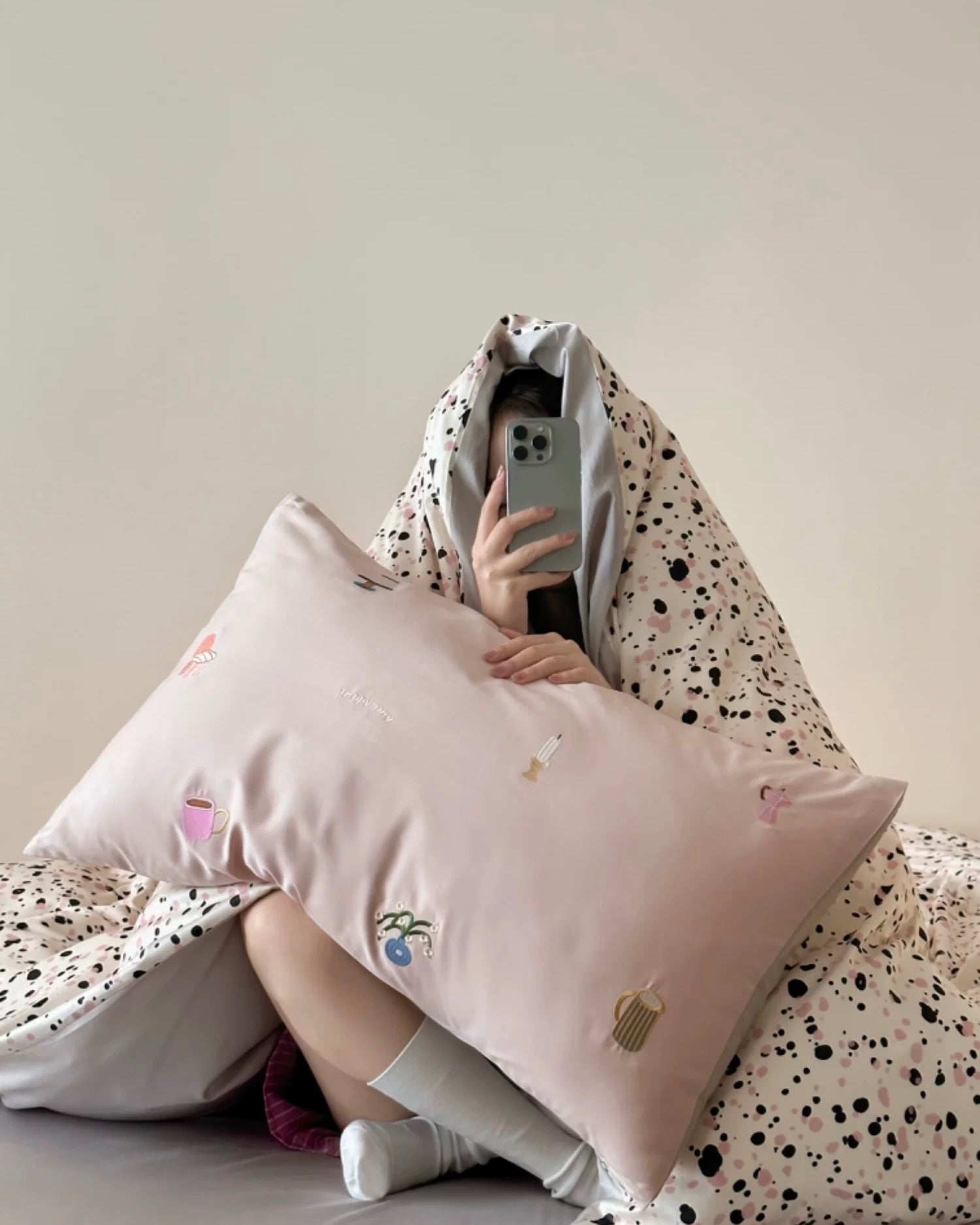 Person sitting on a bed with a pink pillow and a patterned blanket.