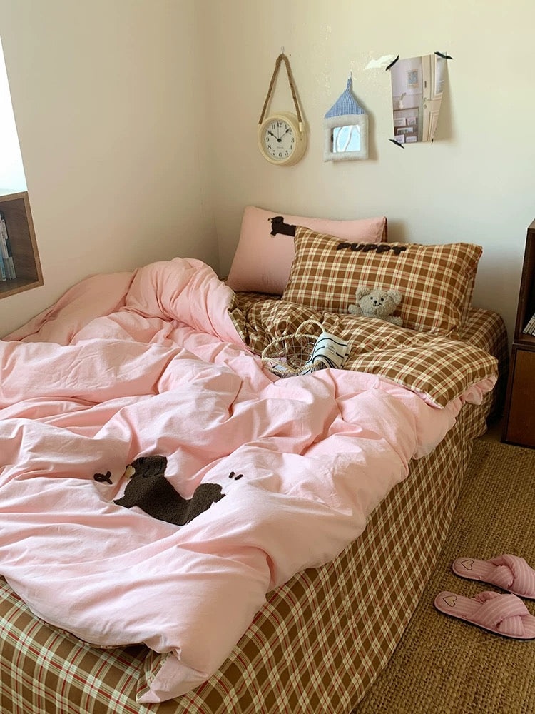 Bed with pink comforter and plaid blanket, pillows, and a clock on the wall.