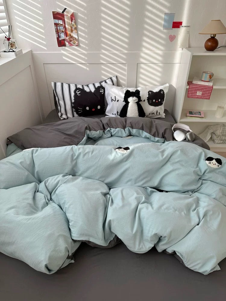 Bed with light blue comforter and cat-themed pillows in a bright room.