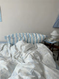 Bed with white and blue floral bedding and striped pillow, nightstand with lamp and bowl in the background.