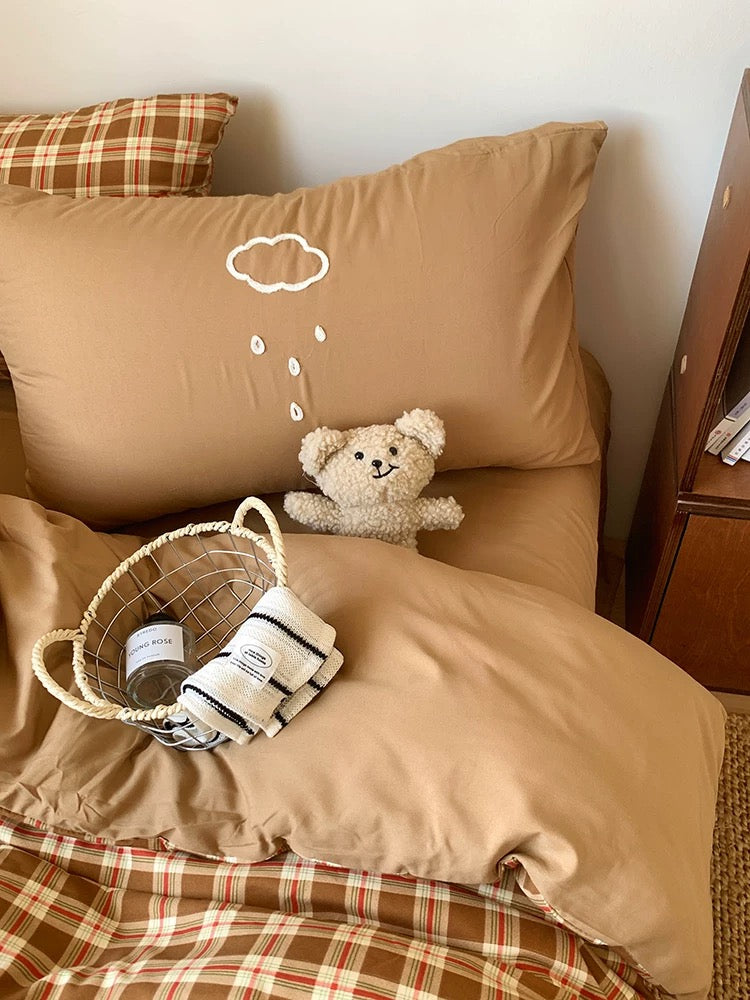 Beige pillow with a teddy bear and basket on a bed with plaid bedding.