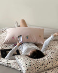 Person lying on a bed with polka dot bedding, using a phone to take a photo of a decorative pillow.