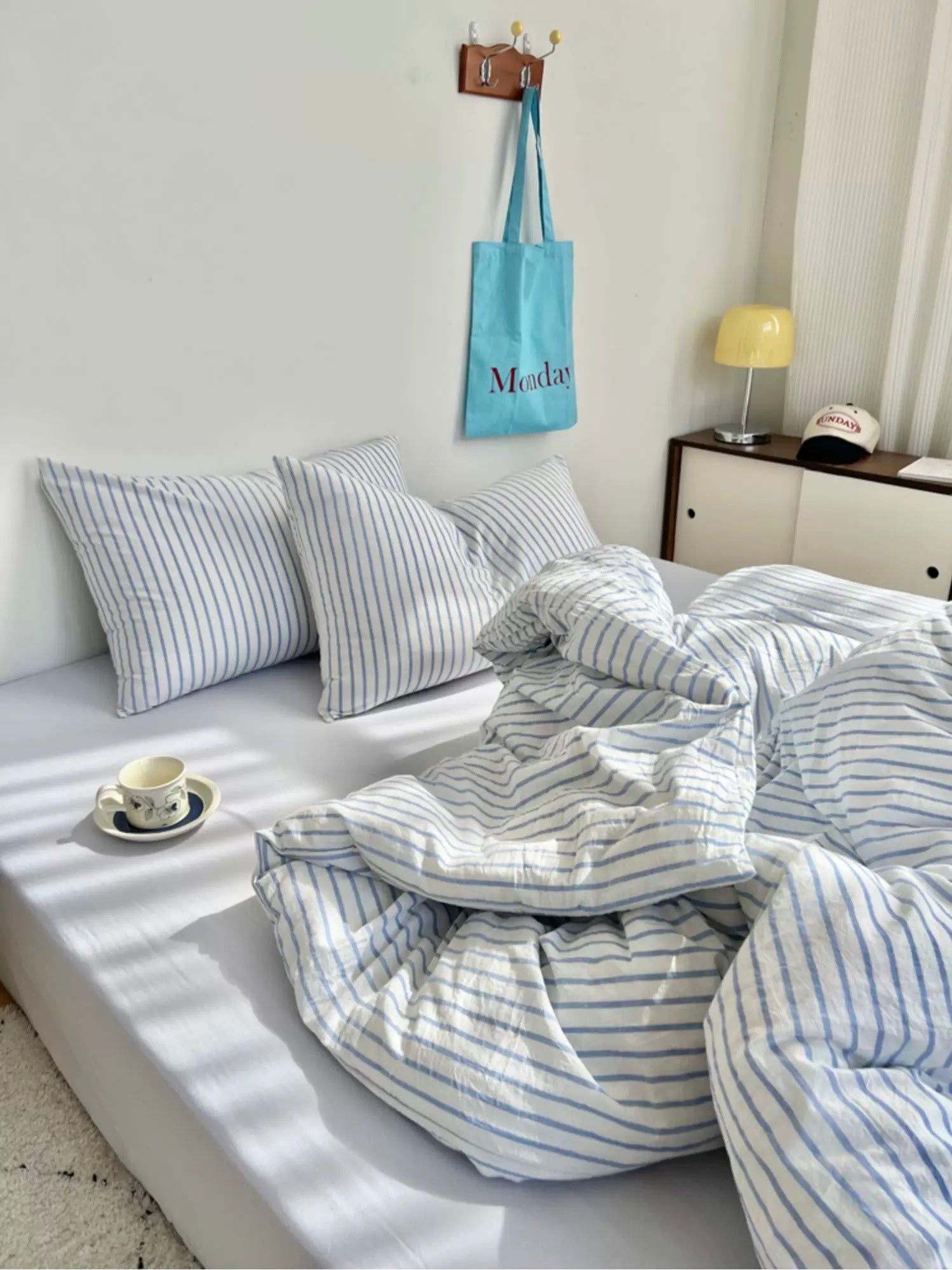 Bed with striped bedding and pillows, light blue bag hanging on wall, and small table with items.