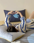 Person lying on a bed with a blue and white patterned blanket, making a heart shape with their hands.