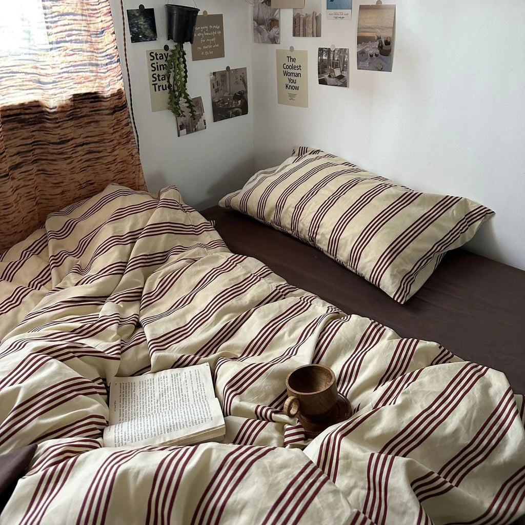Bed with striped bedding and a book, mug, and plant on top in a cozy room.