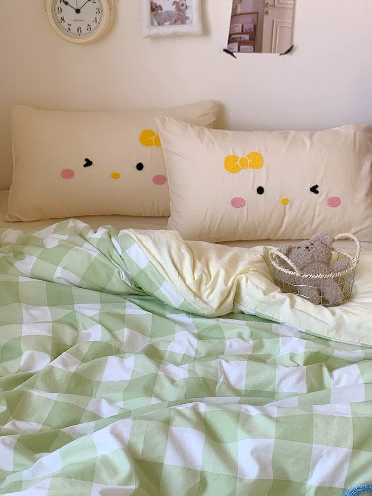 Bed with green checkered blanket, white pillows with smiley face design, and small basket on a light-colored wall background.