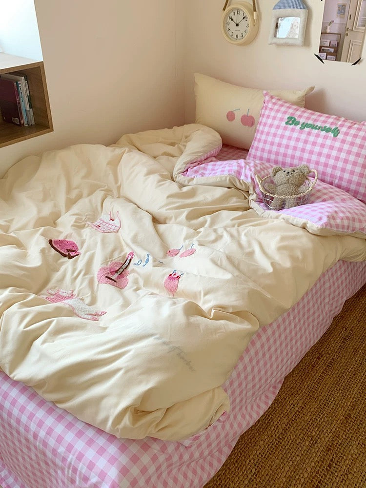 Bed with pink checkered sheets and white comforter, featuring a teddy bear and books on a nightstand.