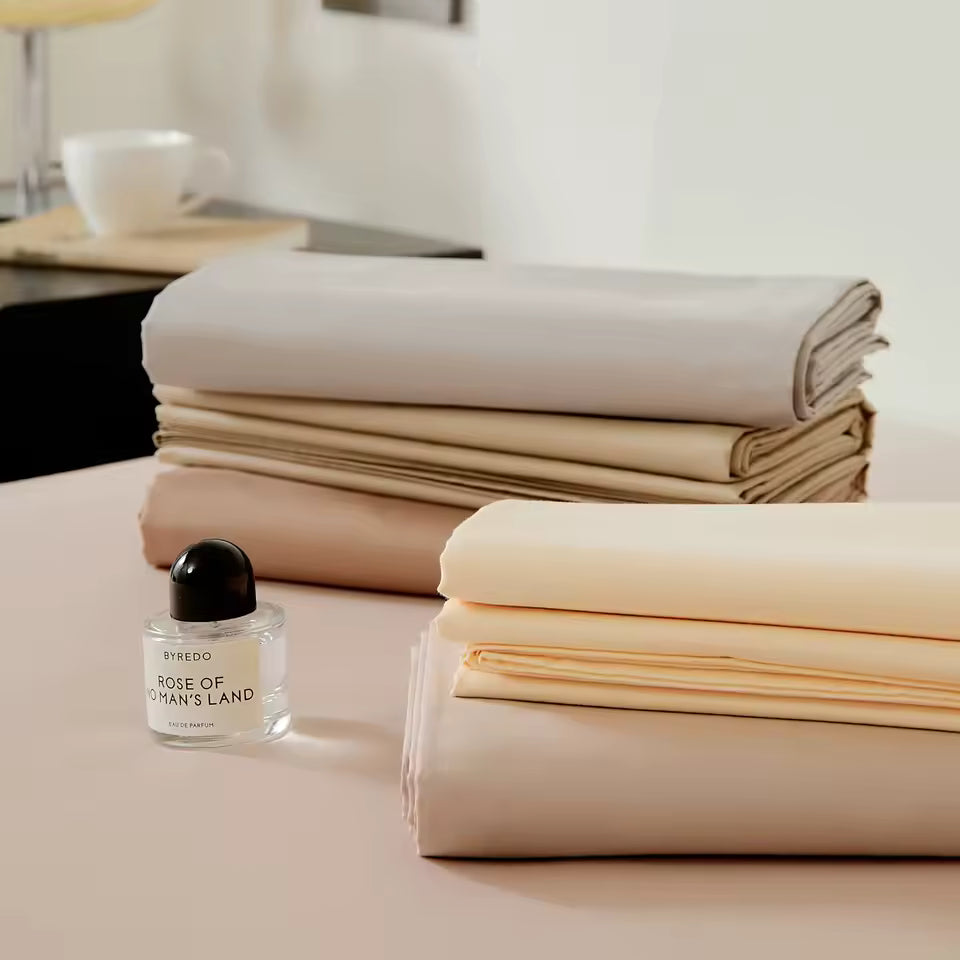 Stack of folded beige sheets on a bed with a small bottle of Byredo perfume.