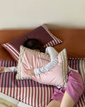 Person lying on a bed with striped bedding and pillows, wearing a pink shirt and white gloves.