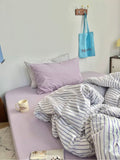 Bed with striped bedding and lavender pillows in a room with a blue bag hanging on the wall.
