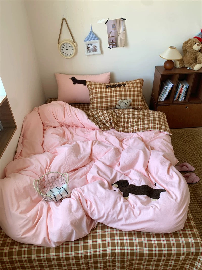 Bed with pink comforter and plaid pillows in a room with a clock and books on a shelf.