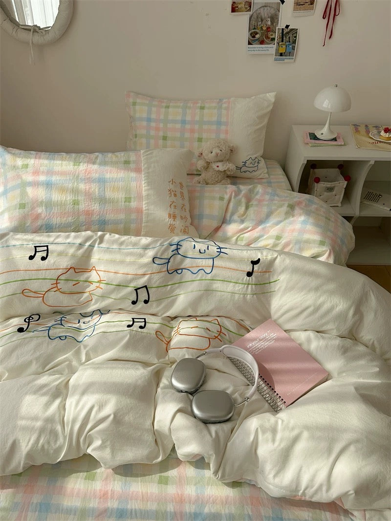Bed with white comforter featuring colorful cat designs, surrounded by pillows and a teddy bear.