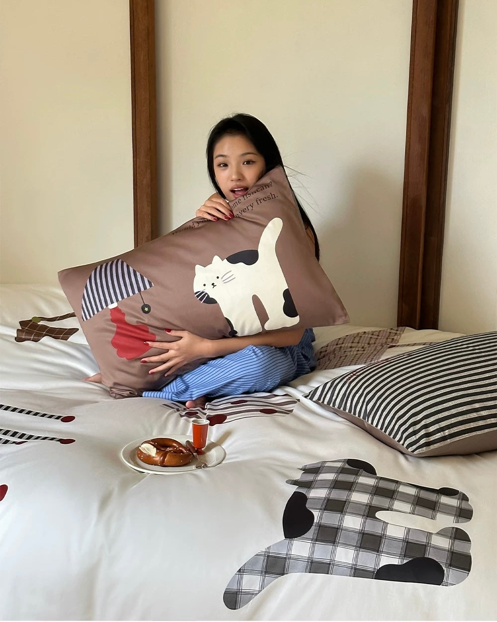 Woman holding a pillow with cartoon cat design on a bed with similar patterned pillows.